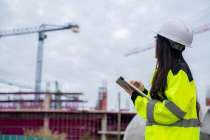 A construction worker in a high-visibility jacket and hard hat inspects a site while holding a tablet.