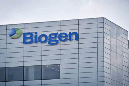 Close-up of the Biogen building exterior, showing the company’s blue and green logo and large blue “Biogen” text on a modern, reflective glass-panelled facade under a clear sky.