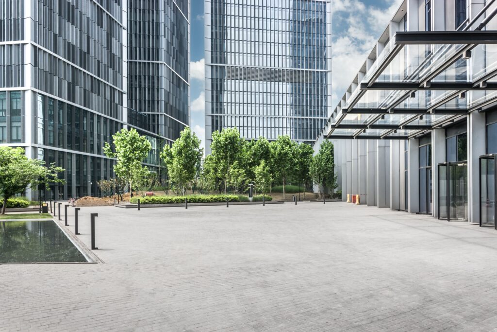 A modern urban courtyard surrounded by tall, glass-fronted office buildings and lush green trees.