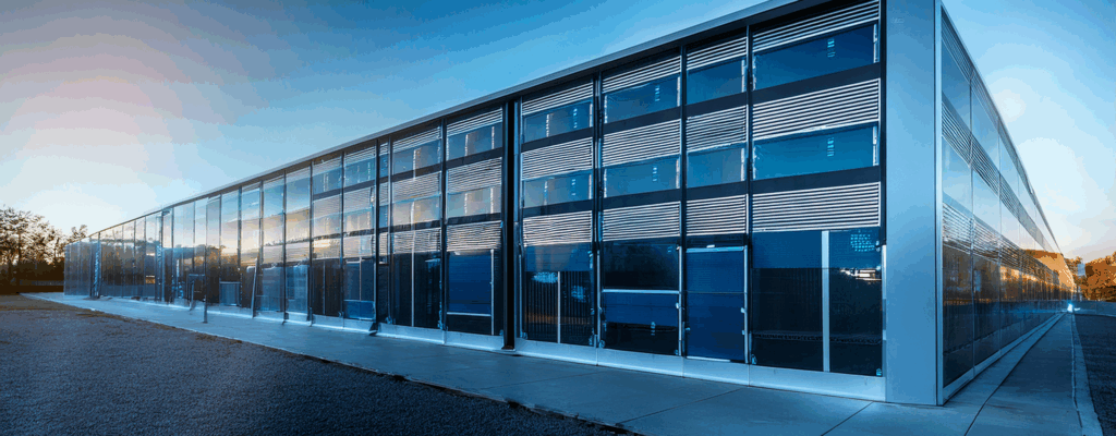 A large, modern data center building with a sleek, gray metal exterior stands under a clear blue sky with a few scattered clouds. The structure has sharp, rectangular lines and is surrounded by an empty asphalt parking lot. One side of the building features large vertical ventilation panels, and the flat roof is lined with multiple cylindrical HVAC units. A single gray metal door with a small canopy is located on the right side, next to a strip of green grass. The scene is brightly lit by daylight, emphasizing the building’s clean and industrial design.
