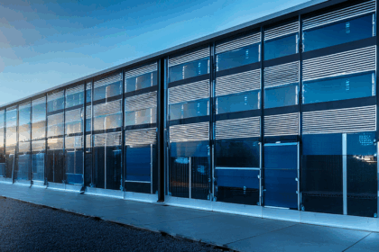 A large, modern data center building with a sleek, gray metal exterior stands under a clear blue sky with a few scattered clouds. The structure has sharp, rectangular lines and is surrounded by an empty asphalt parking lot. One side of the building features large vertical ventilation panels, and the flat roof is lined with multiple cylindrical HVAC units. A single gray metal door with a small canopy is located on the right side, next to a strip of green grass. The scene is brightly lit by daylight, emphasizing the building’s clean and industrial design.