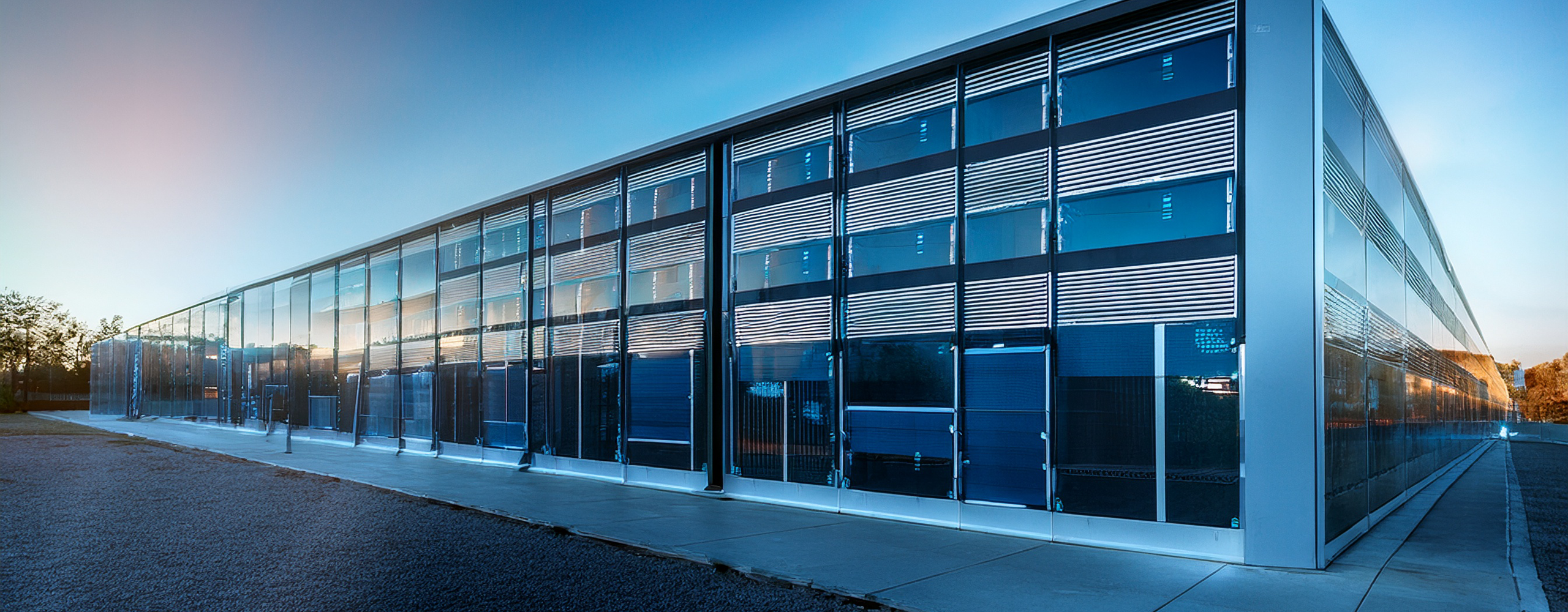 A large, modern data center building with a sleek, gray metal exterior stands under a clear blue sky with a few scattered clouds. The structure has sharp, rectangular lines and is surrounded by an empty asphalt parking lot. One side of the building features large vertical ventilation panels, and the flat roof is lined with multiple cylindrical HVAC units. A single gray metal door with a small canopy is located on the right side, next to a strip of green grass. The scene is brightly lit by daylight, emphasizing the building’s clean and industrial design.