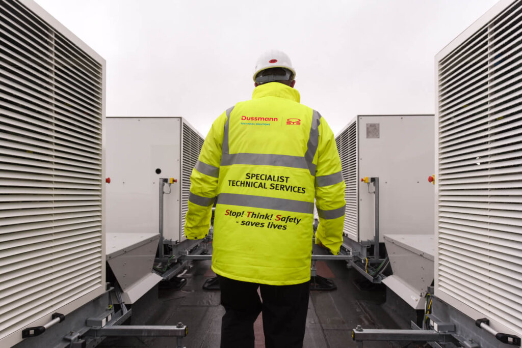 A worker in a high-visibility jacket and hard hat, labeled "Dussmann Specialist Technical Services," stands between large industrial units.