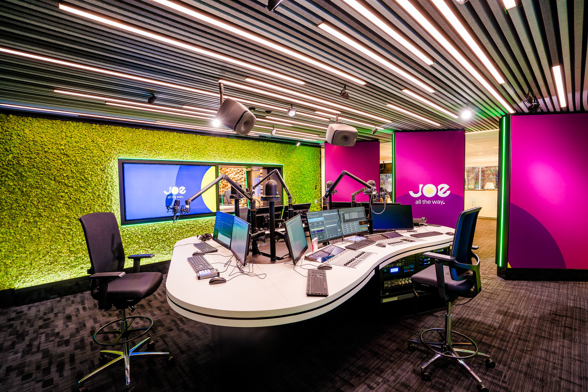 A modern radio studio with vibrant green and purple walls, equipped with multiple screens and microphones.