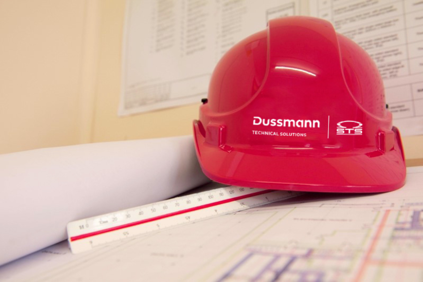 A red hard hat with "Dussmann Technical Solutions" on it rests on blueprints and a ruler.