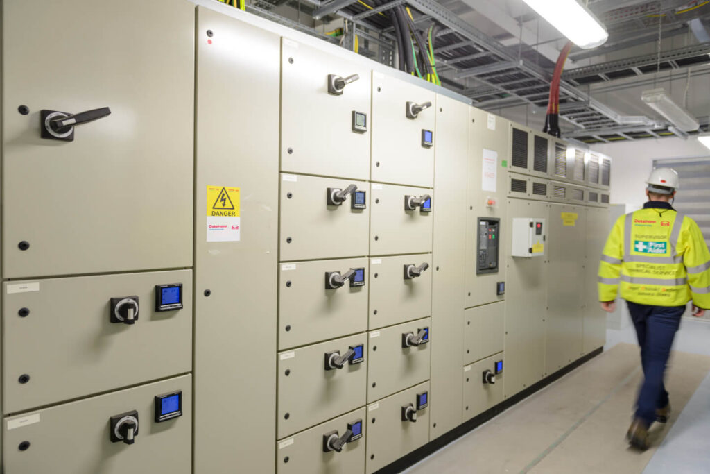 A worker in a high-visibility vest walks past electrical control panels.