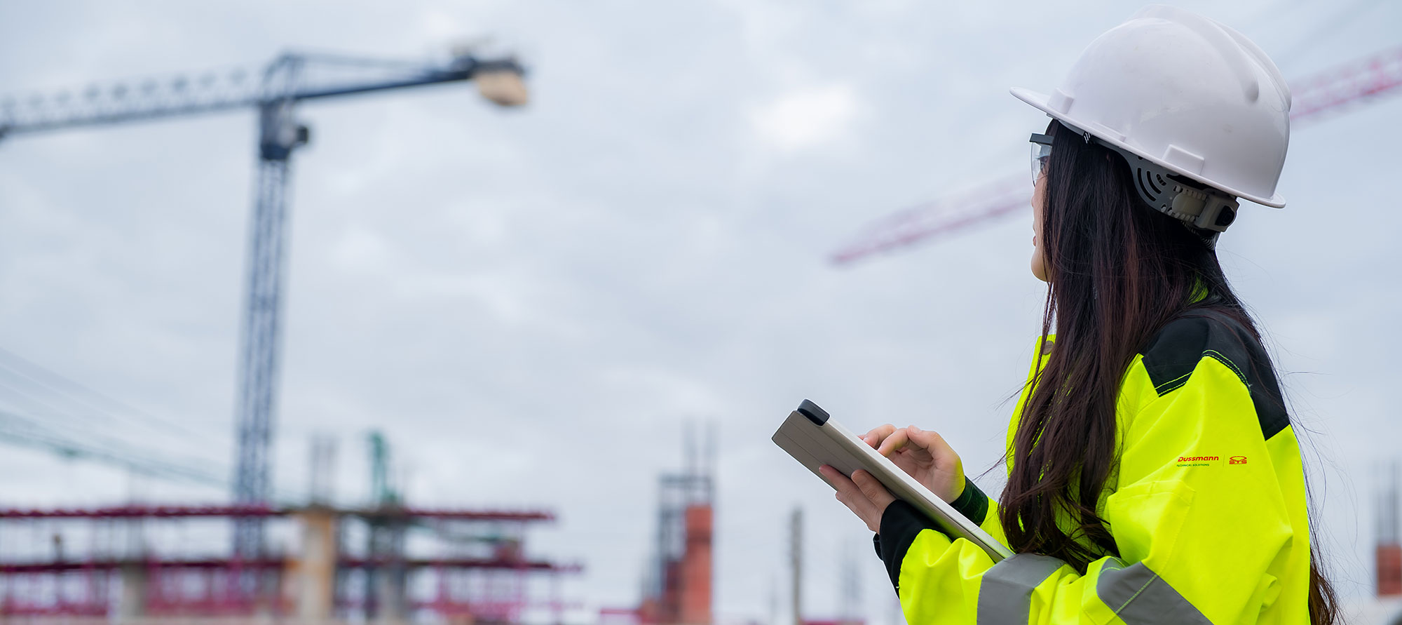 A construction worker in a high-visibility jacket and hard hat inspects a site while holding a tablet.