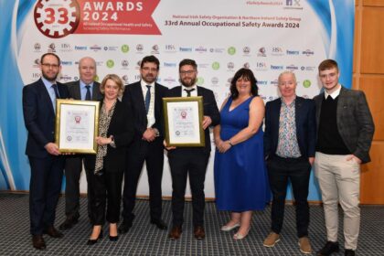 This image features a group of eight people posing at an awards ceremony. The backdrop behind them displays the event name: the "33rd Annual Occupational Safety Awards 2024," organized by the National Irish Safety Organisation and Northern Ireland Safety Group. The design includes a blue and white theme with multiple sponsor logos. Two individuals in the front are holding framed award certificates, smiling proudly. The group is dressed in formal and semi-formal attire, with men wearing suits, blazers, or dress shirts, and a woman in an elegant blue dress. The setting appears to be an official conference or awards hall, with a professional carpet and lighting. Everyone in the group looks happy, likely celebrating their achievement in occupational health and safety.