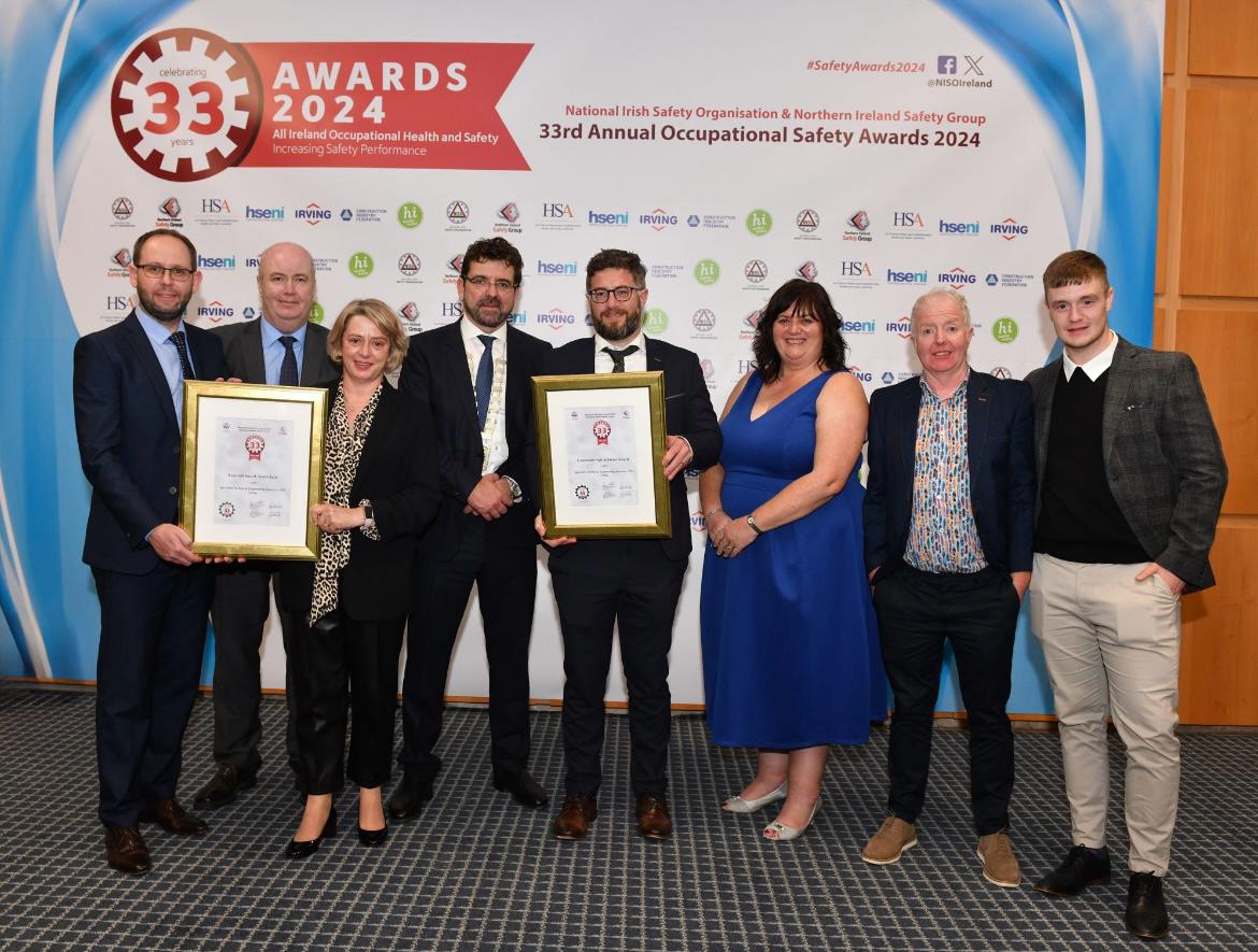 This image features a group of eight people posing at an awards ceremony. The backdrop behind them displays the event name: the "33rd Annual Occupational Safety Awards 2024," organized by the National Irish Safety Organisation and Northern Ireland Safety Group. The design includes a blue and white theme with multiple sponsor logos. Two individuals in the front are holding framed award certificates, smiling proudly. The group is dressed in formal and semi-formal attire, with men wearing suits, blazers, or dress shirts, and a woman in an elegant blue dress. The setting appears to be an official conference or awards hall, with a professional carpet and lighting. Everyone in the group looks happy, likely celebrating their achievement in occupational health and safety.