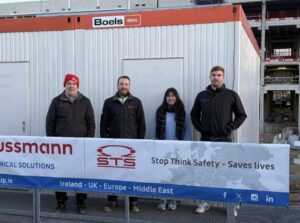 This image features four individuals standing in front of a construction site with a temporary office unit. They are posing behind a safety banner that reads, "Stop Think Safety – Saves Lives," alongside logos for "STS" and "Hussmann Technical Solutions." The banner also highlights the company's presence in Ireland, the UK, Europe, and the Middle East. The group is dressed warmly, with three of them wearing black jackets with company logos, and one person wearing a red beanie. The construction site behind them appears active, with scaffolding and construction materials visible. This seems to be a workplace safety awareness or promotional image emphasizing the company's commitment to safety practices on-site.