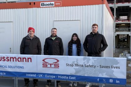 This image features four individuals standing in front of a construction site with a temporary office unit. They are posing behind a safety banner that reads, "Stop Think Safety – Saves Lives," alongside logos for "STS" and "Hussmann Technical Solutions." The banner also highlights the company's presence in Ireland, the UK, Europe, and the Middle East. The group is dressed warmly, with three of them wearing black jackets with company logos, and one person wearing a red beanie. The construction site behind them appears active, with scaffolding and construction materials visible. This seems to be a workplace safety awareness or promotional image emphasizing the company's commitment to safety practices on-site.