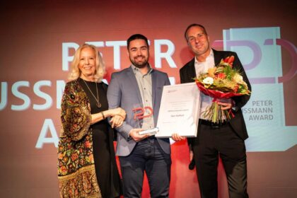 Three people standing on stage at an awards ceremony. The person in the center, a man in a blue blazer, is holding a trophy and a certificate that reads 'Peter Ussmann Award.' To his left, a woman with blonde hair, wearing a patterned shawl, is shaking his hand and smiling. To his right, a man in a black suit is holding a bouquet of flowers. The background features a large screen displaying 'Peter Ussmann Award' in bold text.