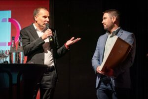 This image captures a moment on stage during an awards ceremony. Two men are engaged in conversation under bright stage lighting. The man on the left is wearing a black suit with a white dress shirt and is holding a microphone while speaking. He gestures with his free hand, emphasizing his words. His facial expression is serious yet engaged. The man on the right is dressed in a light blue blazer and a light-colored shirt. He holds a framed certificate or award against his body and listens attentively, looking at the speaker with a slight smile. A small round table is positioned nearby with bottles of water and empty wine glasses on it. The background features a red-lit screen with partial text and graphics, consistent with an awards event. The lighting highlights the subjects, creating a contrast with the darker background.