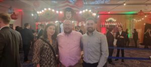 This image captures a lively indoor event with a carnival or circus theme. Three people are posing together in the foreground, smiling at the camera. The person in the middle is a man wearing glasses and a light pink button-up shirt. He has his arms around the other two individuals, a woman on his left and a man on his right. The woman is dressed in a patterned outfit with a crossbody bag, while the man on the right is wearing a light-colored dress shirt. The background is filled with people socializing, dressed in semi-formal attire. There are red and white striped booths, decorative lights, and a clown face above one of the structures, adding to the festive atmosphere. String lights and red lanterns hang from the ceiling, creating a warm and colorful ambiance.