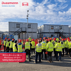 A group of workers wearing high-visibility vests and red safety helmets gathers outdoors around a safety instructor for a Mobile Elevated Work Platform (MEWP) rescue demonstration. The event takes place in front of temporary site office buildings under a blue sky. Branded logos for Dussmann Technical Solutions and STS are visible in the top corners of the image.