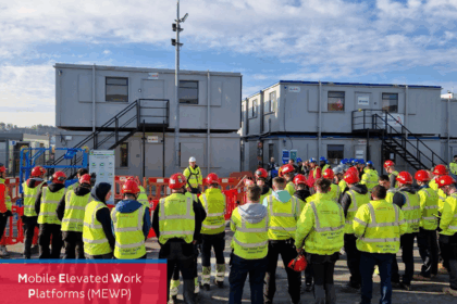 A group of workers wearing high-visibility vests and red safety helmets gathers outdoors around a safety instructor for a Mobile Elevated Work Platform (MEWP) rescue demonstration. The event takes place in front of temporary site office buildings under a blue sky. Branded logos for Dussmann Technical Solutions and STS are visible in the top corners of the image.