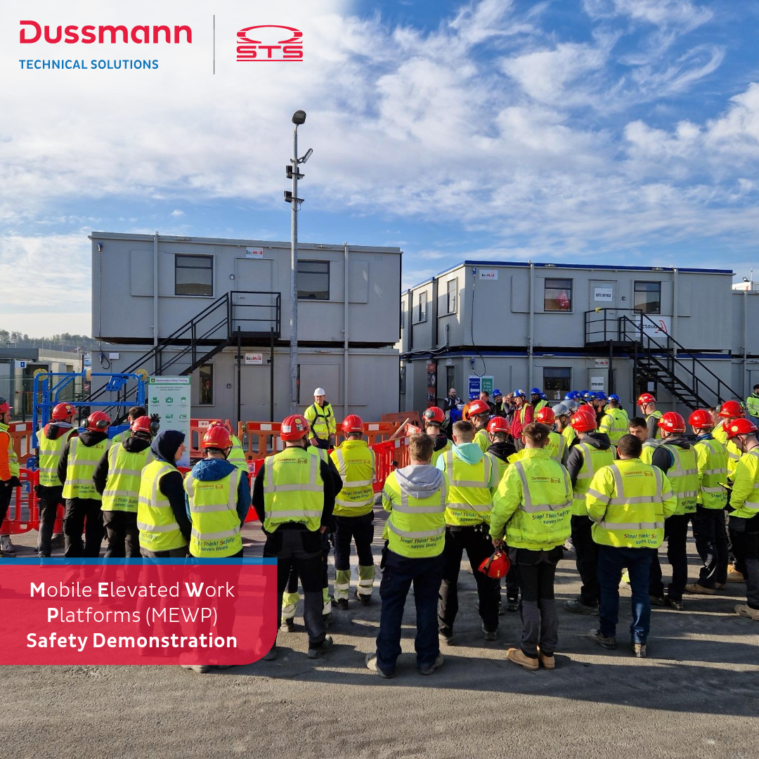 A group of workers wearing high-visibility vests and red safety helmets gathers outdoors around a safety instructor for a Mobile Elevated Work Platform (MEWP) rescue demonstration. The event takes place in front of temporary site office buildings under a blue sky. Branded logos for Dussmann Technical Solutions and STS are visible in the top corners of the image.