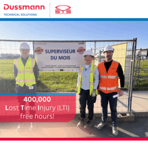 Three construction workers stand in front of a “Supervisor of the Month” sign at a worksite. They wear safety gear and high-vis vests. A banner reads “400,000 Lost Time Injury (LTI) free hours!” with Dussmann Technical Solutions and STS logos above.