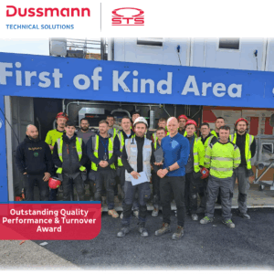 A group of construction workers stands proudly in front of a sign reading "First of Kind Area." Two men at the front hold an award, celebrating their team’s achievement. The banner highlights: "Outstanding Quality Performance & Turnover Award.