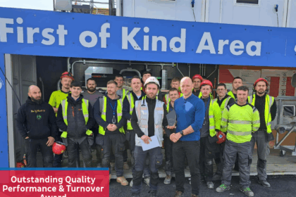 A group of construction workers stands proudly in front of a sign reading "First of Kind Area." Two men at the front hold an award, celebrating their team’s achievement. The banner highlights: "Outstanding Quality Performance & Turnover Award.
