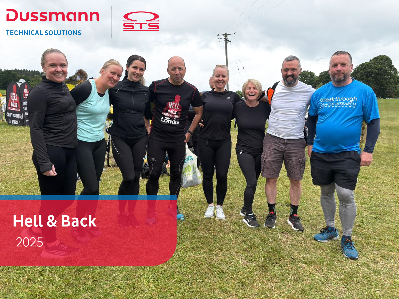 A group of eight smiling people standing together outdoors on grass after completing the 'Hell & Back' challenge in 2025. They're dressed in athletic gear, some muddy from the event. Behind them are event signs and trees under a cloudy sky. The Dussmann Technical Solutions and STS logos are shown at the top left, with a red banner reading 'Hell & Back 2025' at the bottom left.