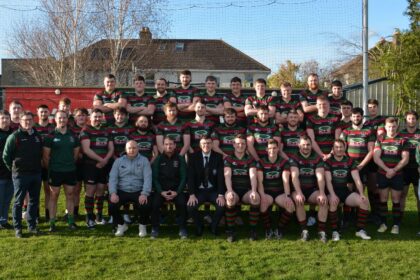 Group photo of the Highfield RFC rugby team and coaching staff posing on a grass field. The players are wearing black, green, and red striped jerseys. The background includes a mural on a wall with “HIGHFIELD” painted on it and a rugby player illustration, with houses and trees behind the fence under a clear blue sky.