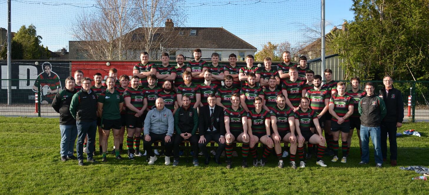 Group photo of the Highfield RFC rugby team and coaching staff posing on a grass field. The players are wearing black, green, and red striped jerseys. The background includes a mural on a wall with “HIGHFIELD” painted on it and a rugby player illustration, with houses and trees behind the fence under a clear blue sky.