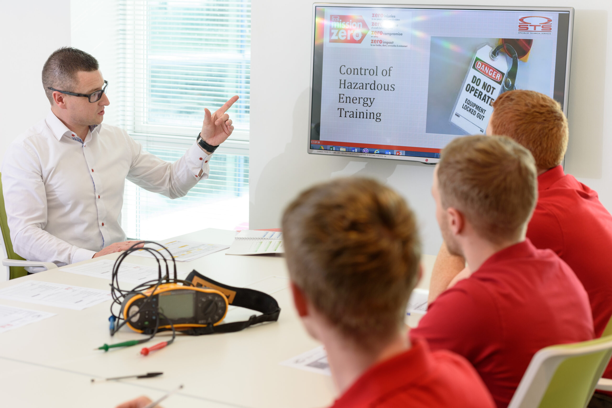 "A safety training session in progress. A man in a white shirt is seated at a table, pointing at a wall-mounted screen displaying a presentation titled 'Control of Hazardous Energy Training.' The screen also shows an image of a danger tag reading 'DO NOT OPERATE – Equipment Locked Out.' Three men in red shirts are seated and attentively facing the presenter. On the table are printed documents and an electrical testing device with cables."