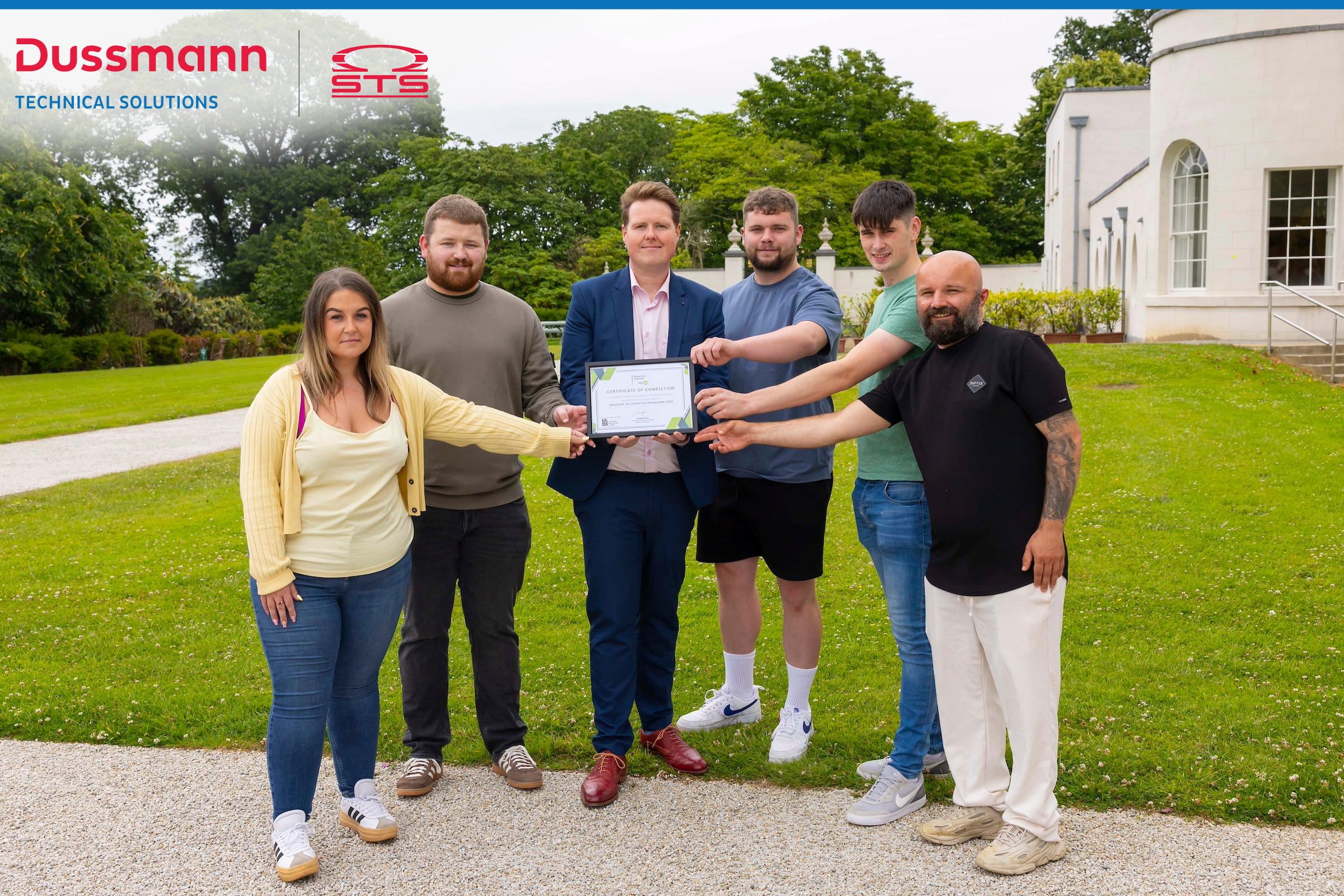 Six people stand outdoors on a lawn in front of a white building, smiling and holding a certificate together. The person in the center, dressed in a navy suit, holds the certificate while the others extend their arms to touch it, celebrating a shared achievement. The Dussmann Technical Solutions and STS logos appear at the top left of the image. Trees and a path provide a scenic background.