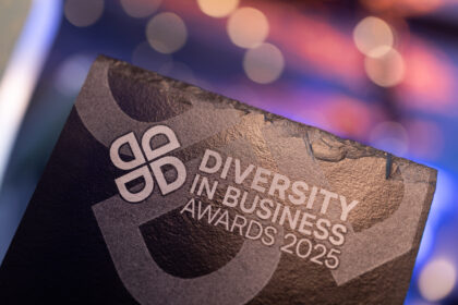 A close-up image of a black award plaque with the text 'Diversity in Business Awards 2025' engraved on it. The background is blurred with colorful bokeh lights.