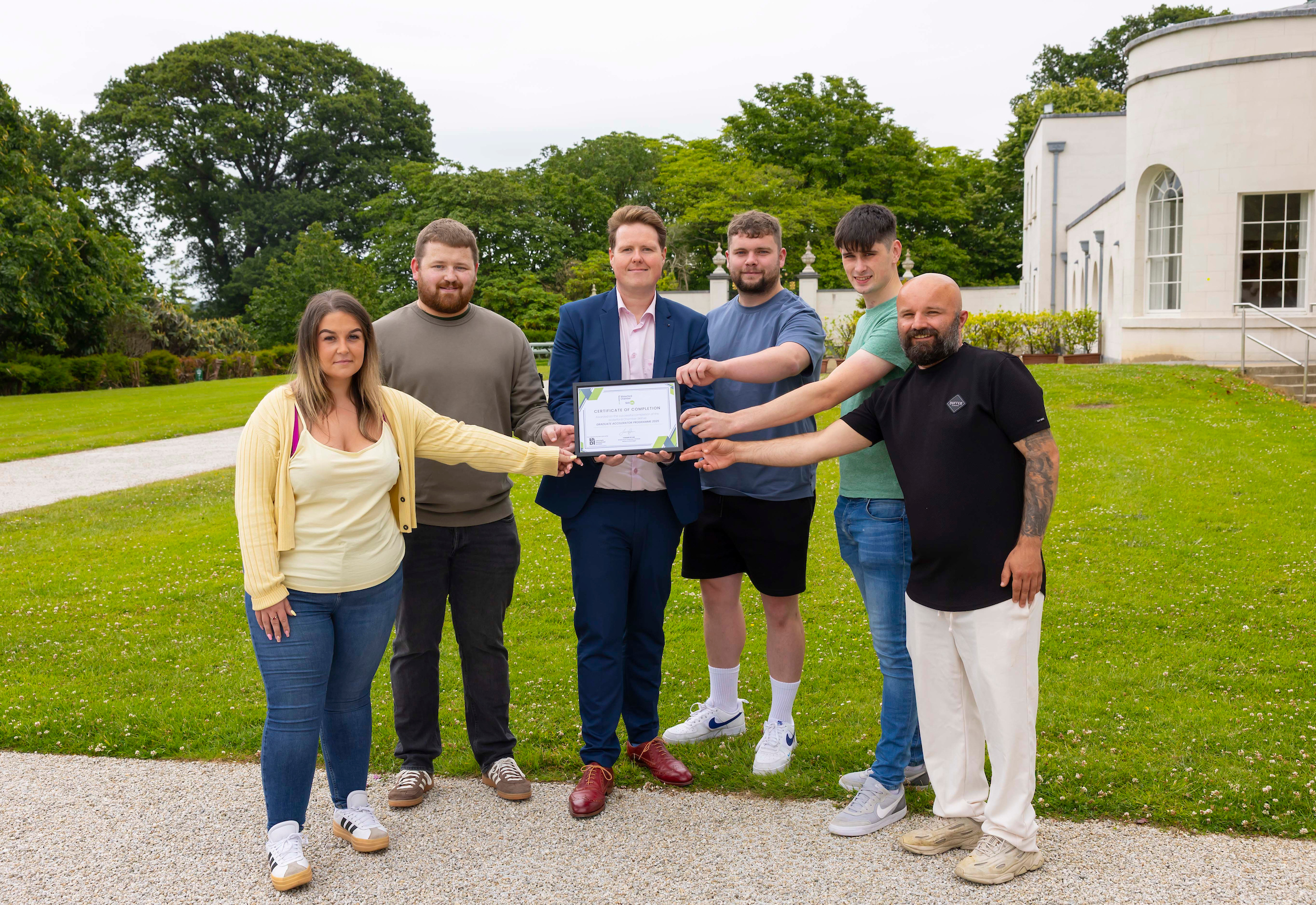 Six people stand outdoors on a lawn in front of a white building, smiling and holding a certificate together. The person in the center, dressed in a navy suit, holds the certificate while the others extend their arms to touch it, celebrating a shared achievement. The Dussmann Technical Solutions and STS logos appear at the top left of the image. Trees and a path provide a scenic background.