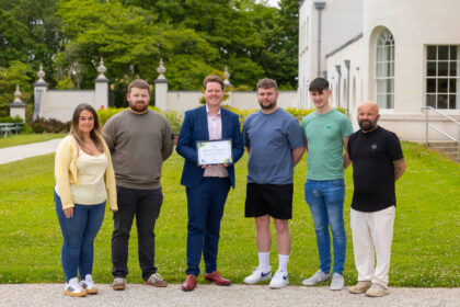 A group of six people stand together outside on a lawn, posing for a photo in front of a white building with large windows. The person in the center, wearing a navy blue suit, holds a certificate of completion. The group appears to be part of a professional or educational achievement event, possibly a graduation or training completion. Trees and manicured hedges provide a green, scenic background.