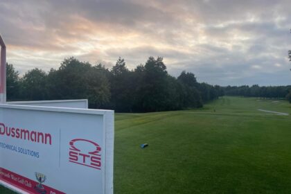 A serene golf course scene with a lush green fairway stretching into the distance, flanked by tall trees on both sides. The sky is partly cloudy with soft sunlight filtering through. In the foreground, a large event sign displays the logos and names 'Dussmann Technical Solutions' and 'STS', indicating sponsorship for a Pro-Am golf event