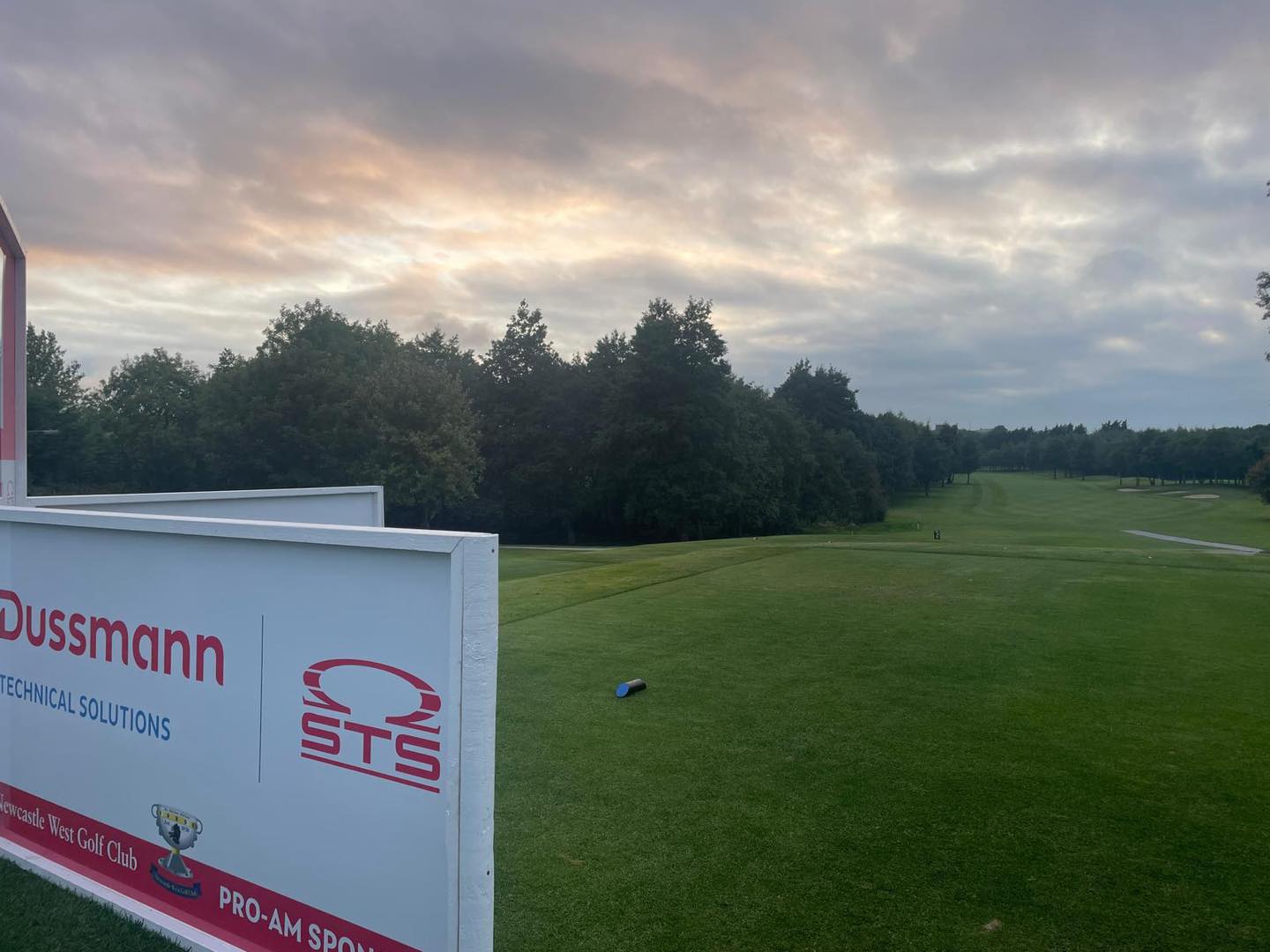 A serene golf course scene with a lush green fairway stretching into the distance, flanked by tall trees on both sides. The sky is partly cloudy with soft sunlight filtering through. In the foreground, a large event sign displays the logos and names 'Dussmann Technical Solutions' and 'STS', indicating sponsorship for a Pro-Am golf event